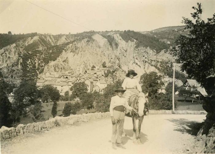 [Homme guidant une mule bâtée sur laquelle est assise une jeune femme, sur la route de Péone, 2e quart du 20e siècle.]